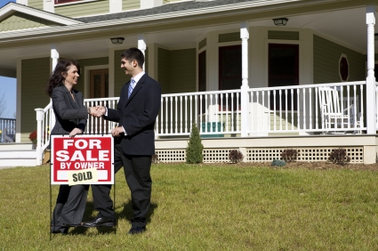 Two people shake hands as they place a "for sale" sign in front of a house. This could be two realtors who have sold the house or a realtor and buyers.